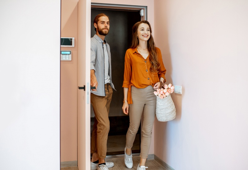 A smiling couple enters a modern apartment through a secure door equipped with a keypad lock on the wall. This image highlights seamless resident access solutions, showcasing how RemoteLock enhances multifamily property management and improves the resident experience.