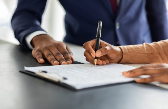 This image depicts a close-up of two people interacting over a document on a table. One individual is holding a pen and appears to be signing the document, while the other person places a hand near the document, possibly guiding or supervising. Both individuals are dressed in professional attire, suggesting a formal or business setting.