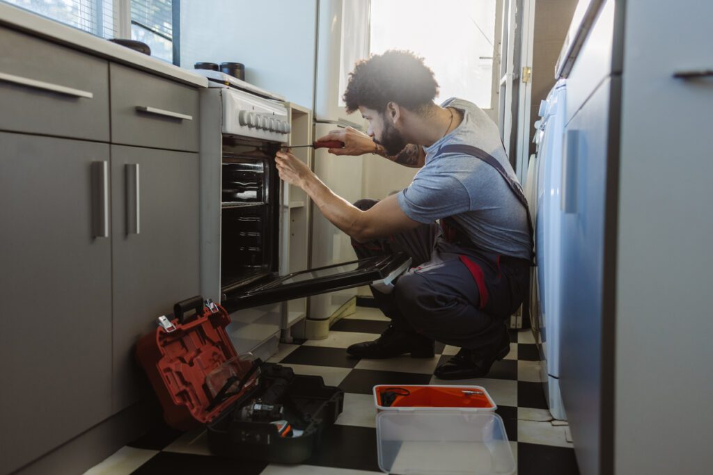 This image shows a technician or handyman repairing an oven in a modern kitchen. The individual is kneeling on the floor, using a screwdriver to work on the appliance. Nearby, a red toolbox and other tools are visible, indicating the repair process. The kitchen features a clean, organized space with checkered flooring and natural light streaming in through a window or door.