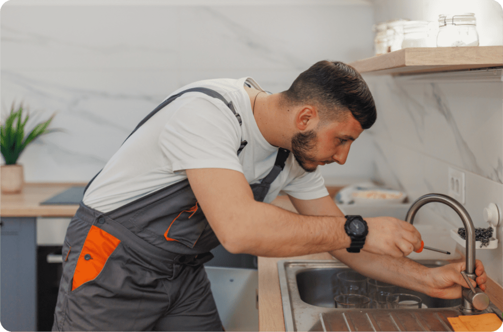 A maintenance worker in overalls repairing a kitchen faucet in a modern, well-lit space. This image highlights the importance of streamlined access for maintenance teams, showcasing how RemoteLock enables efficient property management by providing secure, scheduled access to service providers.