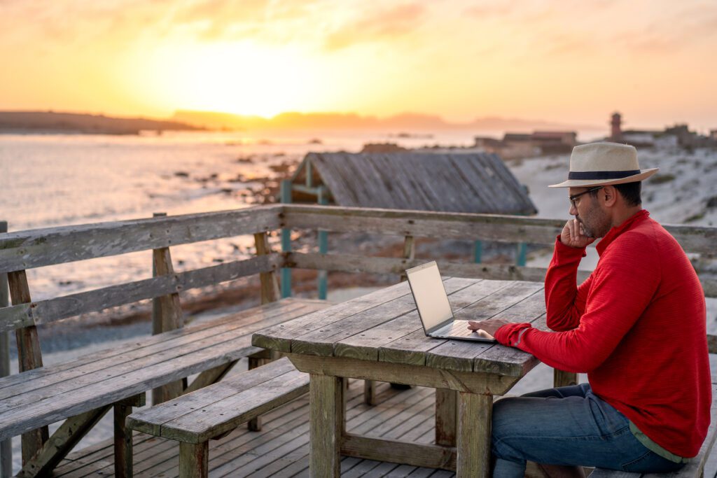 The image shows a man sitting at a wooden picnic table outdoors, working on a laptop. He is wearing a red sweater, a beige hat, and glasses, and appears focused on his work. The setting is by the seaside, with a stunning sunset in the background casting warm golden hues over the scene. Rustic wooden structures and a sandy shoreline are visible nearby, adding to the serene and remote atmosphere of the location.