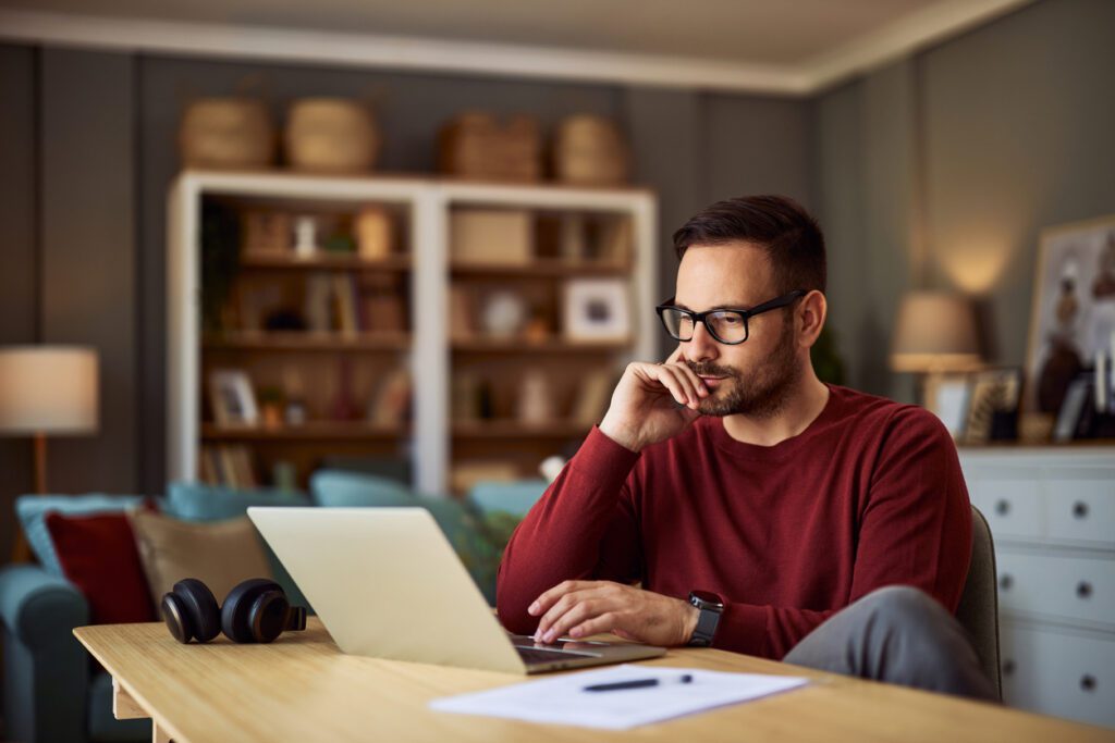 The image shows a man working on a laptop in a cozy, well-lit home office or living room. He is wearing glasses and a red sweater, with a thoughtful expression as he focuses on his work. A pair of headphones rests on the wooden desk, alongside a piece of paper and a pen. The background features shelves with decorative items, books, and wicker baskets, as well as a sofa with colorful cushions, creating a comfortable and organized environment.